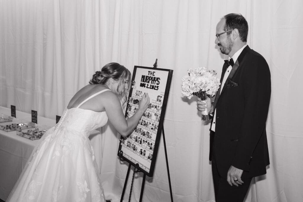 Bride signing a LEGO guest book while the groom watches during a wedding reception at University Club of MSU in Lansing, Michigan