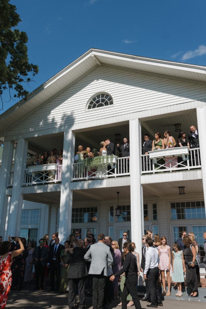 Bride and groom sharing a kiss on the balcony at Walden Woods in Michigan, surrounded by their wedding party and family as guests gather below after their June ceremony.