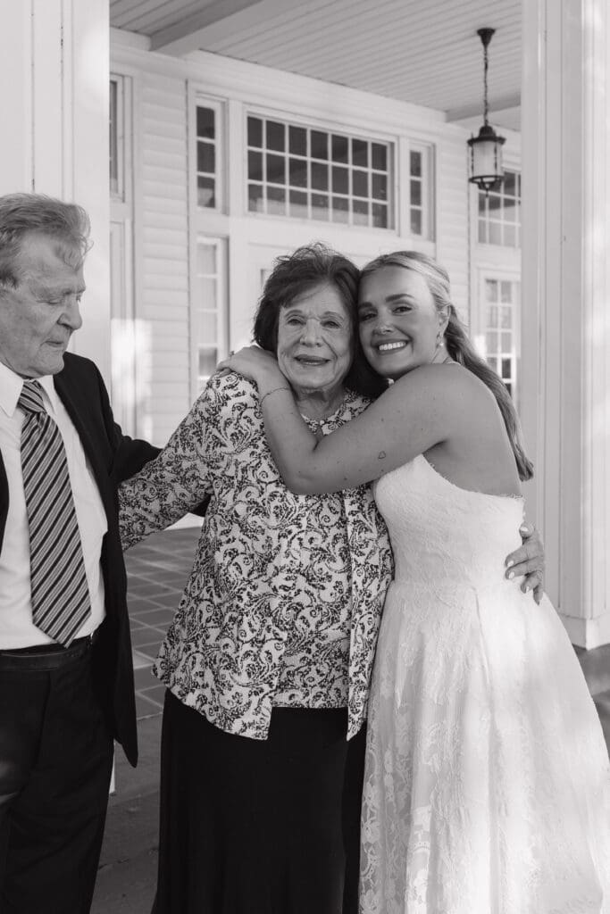 Black and white candid wedding photo of a bride hugging her grandmother outside Walden Woods during a June wedding in Michigan, capturing a heartfelt family moment with her grandfather standing beside them.