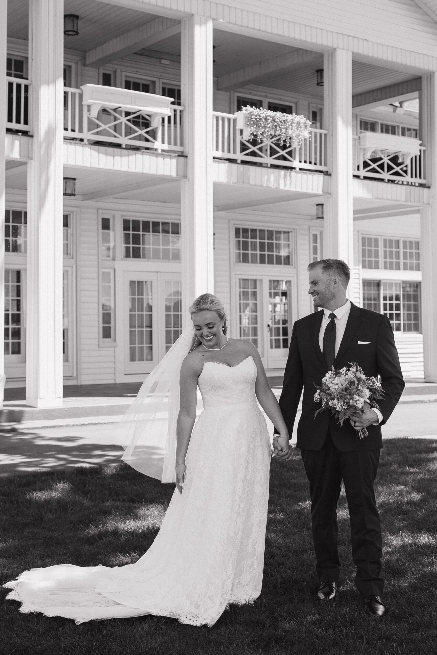 Black and white candid wedding portrait of a bride smiling as she gathers her wedding dress while standing hand in hand with the groom outside Walden Woods in Michigan.