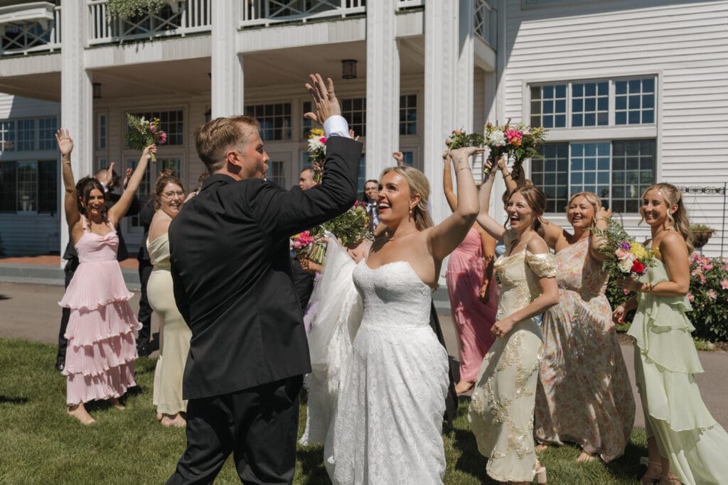 Bride and groom high-fiving during bridal party portraits outside Walden Woods on a sunny June wedding day, surrounded by bridesmaids and groomsmen celebrating with colorful bouquets in Michigan.
