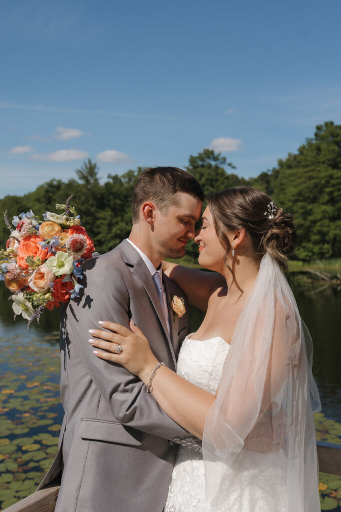 Bride and groom embracing during wedding portraits at Waltman Lake Lodge in West Michigan, standing by the lake with colorful summer flowers, lush greenery, and natural light capturing a candid, romantic moment.