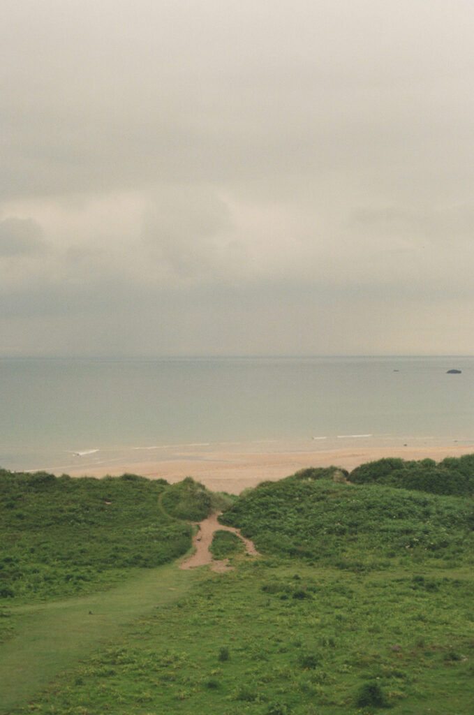 Elevated view of Whitepark Bay Beach along Northern Ireland's Causeway Coast, featuring a winding path through grassy dunes leading to a wide sandy beach and calm Atlantic waters, captured on 35mm film beneath an overcast sky.