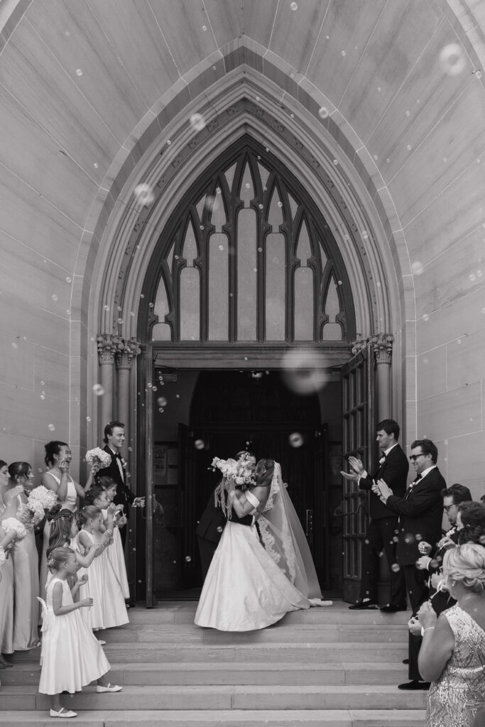 Bride and groom kiss outside St. Andrew's Cathedral in Grand Rapids, Michigan during a bubble exit as family and wedding guests celebrate on the cathedral steps.