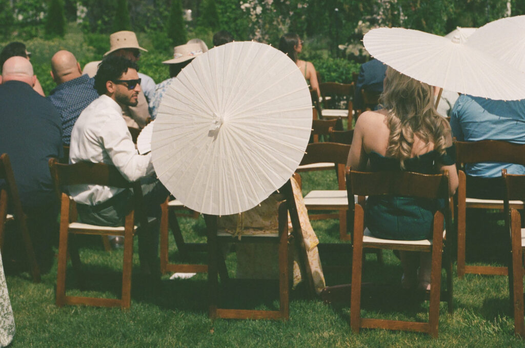 35mm film photograph of wedding guests seated outdoors holding white parasols during a sunny summer ceremony at The Millhaven, capturing the relaxed atmosphere and timeless film aesthetic.