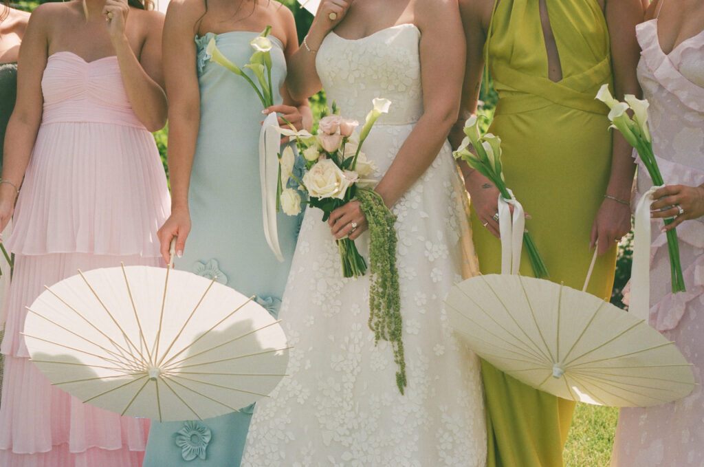 35mm film wedding photo of a bride and bridesmaids holding white parasols and calla lily bouquets during an outdoor summer wedding at The Millhaven, with pastel dresses and natural candid moments captured on film.